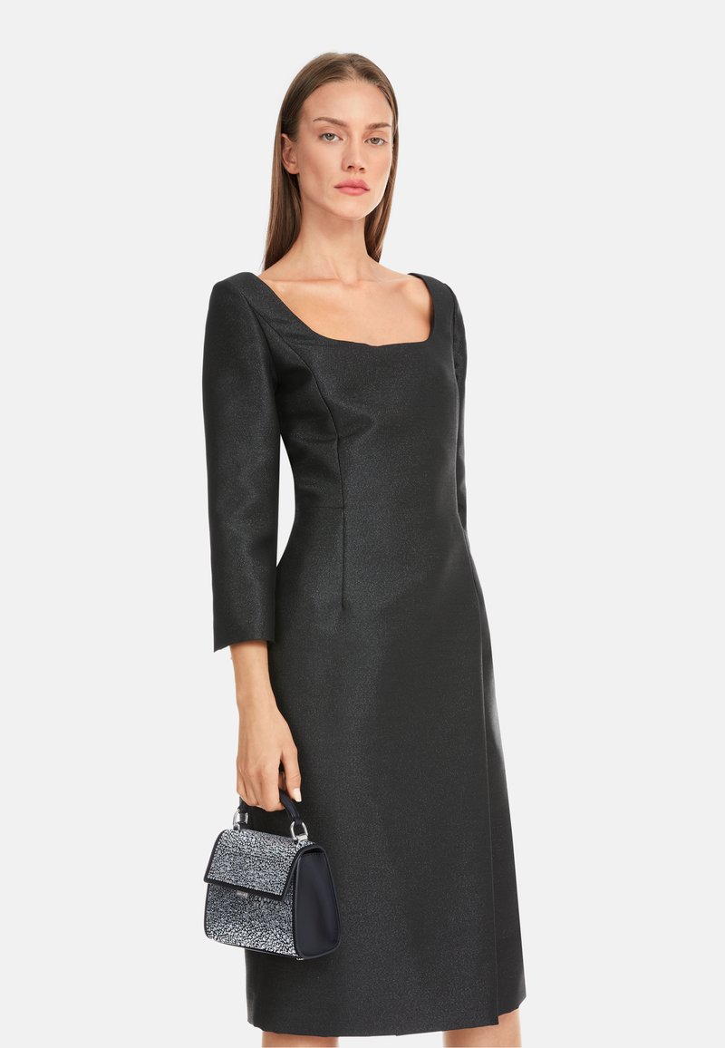 Schwarzes, tailliertes Kleid mit karreeförmigem Ausschnitt und Dreiviertelärmeln. Begleitet von einer kleinen, strukturierten Handtasche in Marineblau und Silber.