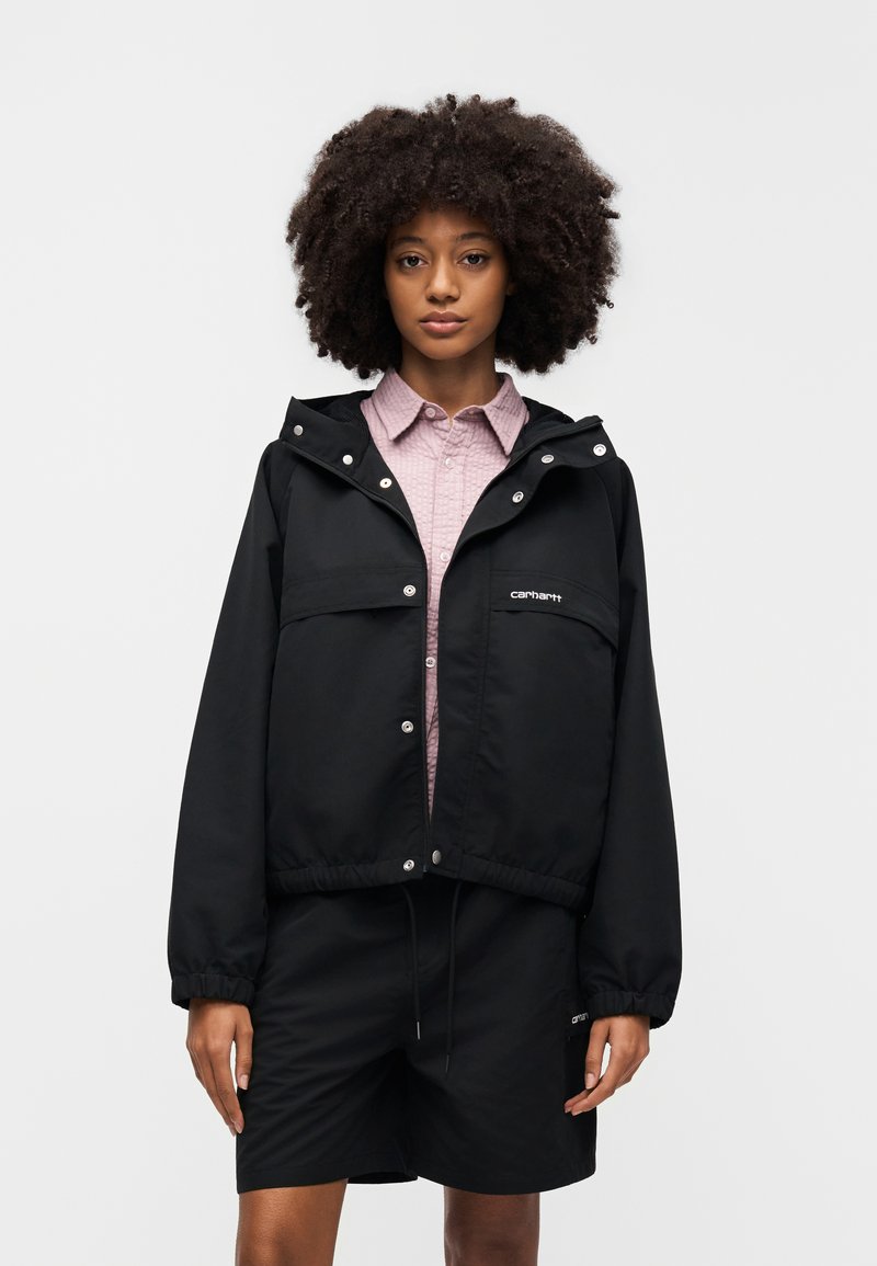 Giacca nera con cappuccio con chiusure a scatto, tasche frontali, orlo elastico e coulisse. Indossata sopra una camicia rosa chiaro con colletto.