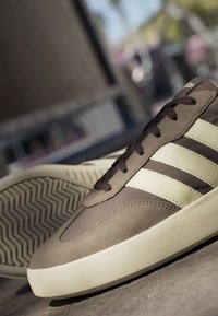Bruine en beige Adidas sneakers met drie witte strepen, bruine veters en een gestructureerde zool schuin op een betonnen oppervlak buiten.