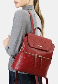 Zaino in pelle rossa con motivo a logo embossato. Presenta un classico coperchio superiore e una tasca frontale con zip. Spallacci regolabili e superficie liscia.