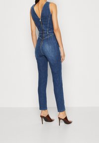 Mono de mezclilla con un escote en V profundo, diseño ajustado y piernas entalladas. Presenta una cremallera en el centro y detalles de costura sutiles. Usado con zapatillas de tacón.