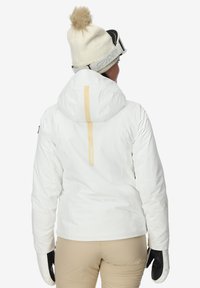 Persoon in een witte skijas, beige broek, witte pomponmuts, zwart-witte skihandschoenen en skibril met 'Spyder' merk.