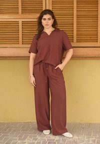 Femme aux longs cheveux foncés portant un haut marron ample et un pantalon, debout avec une main dans la poche contre un volet en bois et un mur jaune.