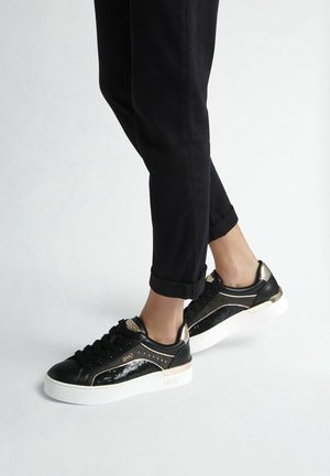 Person iført sorte bukser og sorte sneakers med hvide såler og gyldne detaljer, stående med den ene fod let løftet.