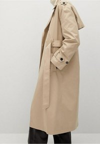 Beżowy trencz z paskiem w talii, z guzikami na mankietach i epoletami. Wykonany z gładkiego materiału, o prostym kroju, sięgającym do połowy łydki.