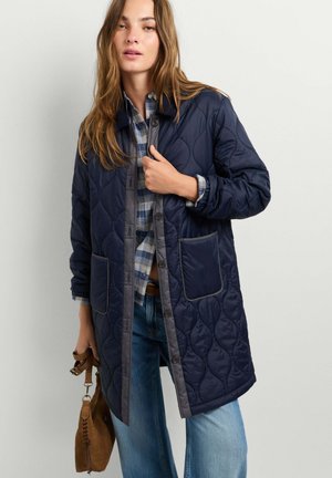 Femme portant un manteau matelassé bleu marine par-dessus une chemise à carreaux et un jean, tenant un sac à main en daim marron à ses côtés.