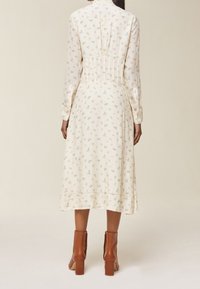 Kvinna i en krämfärgad blommig midiklänning med långa ärmar och bruna ankelstövlar, sedd bakifrån mot en enfärgad bakgrund.