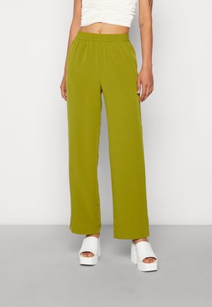 Person iført olivengrønne bukser med vide ben, hvid cropped top og hvide platform sandaletter med åben tå, stående på en grå gulv.