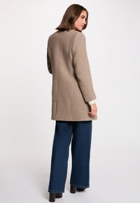 Abrigo de lana beige con corte recto, abertura trasera y detalles de costura visibles, combinado con pantalones de mezclilla de pierna ancha y botas de tacón cuadrado.