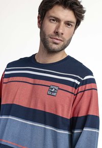 Camisa de manga larga a rayas en azul marino, rojo y azul claro. Cuenta con un pequeño parche con logo en el pecho y un cuello redondo. Textura de tela suave.