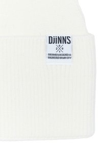 Căciulă tricotată albă cu model rib și etichetă cusută cu "Djinns" și text despre design și inginerie, în negru pe un fundal alb.