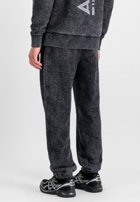 Personne portant un pantalon de jogging noir délavé, un sweat-shirt assorti et des chaussures de sport noires, se tenant devant un fond uni.