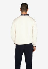 Sudadera ligeramente crema con cuello redondo y puños acanalados, superpuesta a una camisa de cuadros oscuros con cuello. Combinada con pantalones azul marino oscuros.