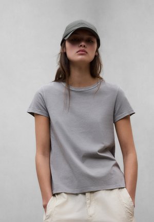 Ung person iført en grå kasket, grå kortærmet t-shirt og lysebeige bukser, stående med hænderne i lommerne foran en ensfarvet baggrund.