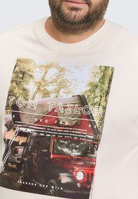 Beige t-shirt met een fotografische print van een tent en een rode voertuig. Witte tekst bevat "TOM TAILOR" en "OMARM DE NATUUR."