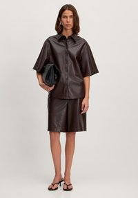 Braunes Lederhemd mit spitzem Kragen, kurzen Ärmeln und Knopfleiste, kombiniert mit passenden Shorts und einer schwarzen Clutch. Schwarze Sandalen.