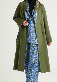 Manteau surdimensionné vert olive à double boutonnage, porté par-dessus un costume bleu à motif floral. Tissu lisse avec une forme structurée.
