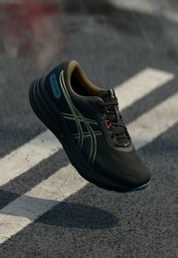 Μαύρο και καφέ αθλητικό παπούτσι ASICS με ετικέτα Gore-Tex, στον αέρα πάνω από γκρι άσφαλτο με λευκές οδικές σημάνσεις.