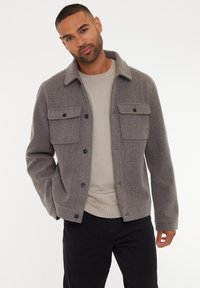 Grå uldblandingsjakke med krave, to brystlommer og knaplukninger. Båret over en beige sweater med ribbet kant.