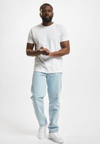 White short-sleeve t-shirt, light blue jeans, white sneakers. Model standing, hands clasped, neutral background.