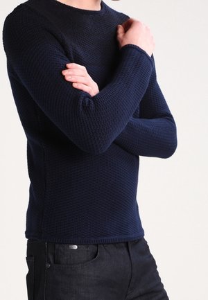 Marineblauer strukturierter Strickpullover mit langen Ärmeln und Rundhalsausschnitt, kombiniert mit schwarzen Denim-Jeans mit einem dezenten Logodetail.