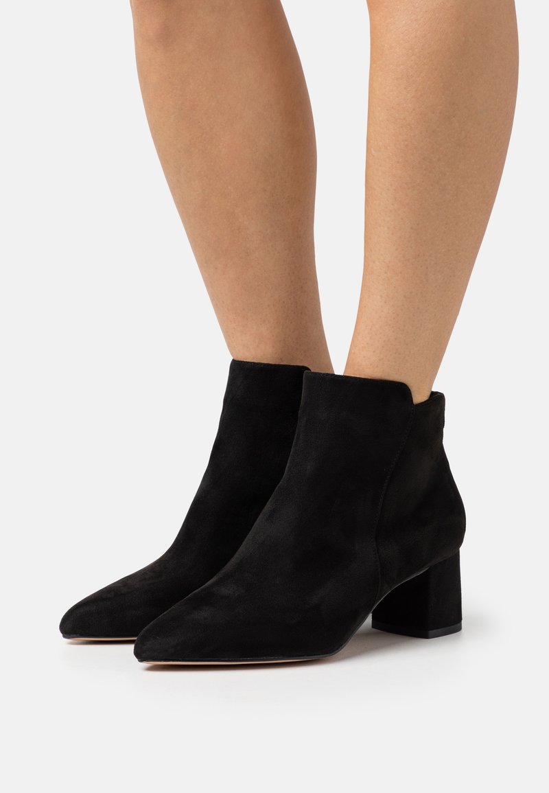 Jambes portant des bottines en daim noir à bouts pointus et talons carrés sur un fond clair uni.