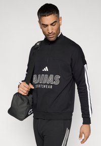 Μαύρο φούτερ Adidas με ψηλό γιακά, λευκό λογότυπο και κάθετες λευκές ρίγες στα μανίκια. Το μοντέλο κρατά μια μαύρη τσάντα.