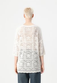 Pull en tricot blanc avec un motif géométrique ajouré, des manches trois-quarts et une coupe décontractée. Le tissu texturé donne une sensation légère.