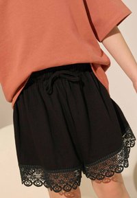 Schwarze Shorts aus strukturiertem Gewebe mit einem elastischen Bund und Spitzenbesatz am Saum. Kombiniert mit einem lockeren, korallenfarbigen Oberteil.