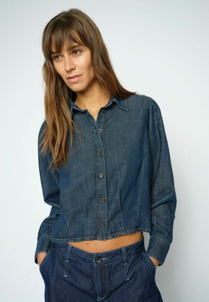 Vrouw met lang bruin haar die een cropped donker denim blouse met knopen draagt en bijpassende donkere denim broek met handen in de zakken.