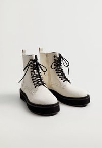 Botas de tobillo blancas con cordones, suelas y cordones negros, que cuentan con cremalleras laterales y tiradores, sobre un fondo liso.