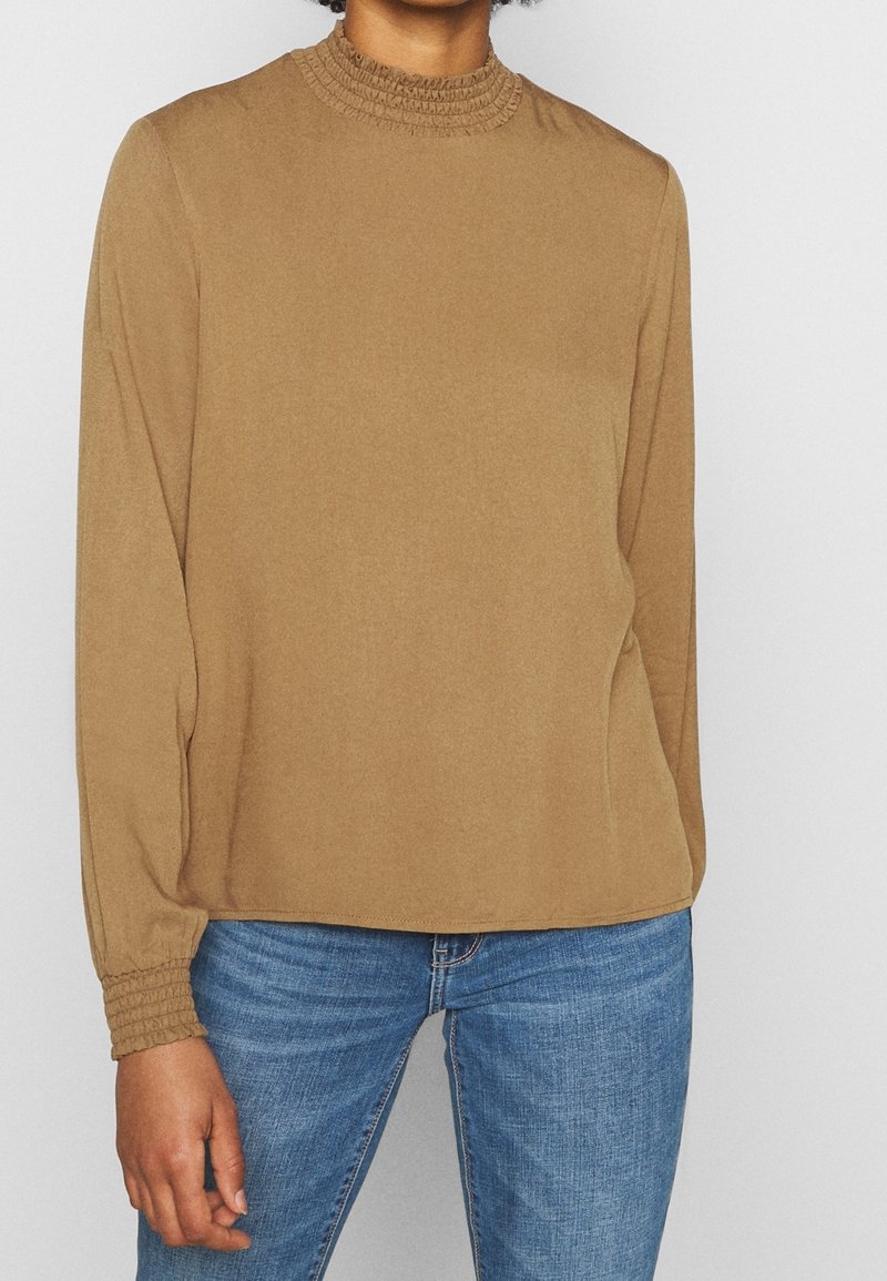Top a maniche lunghe color beige con un collo alto arricciato, vestibilità rilassata e polsini elasticizzati. Abbinato a jeans blu, tessuto morbido.