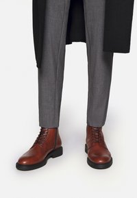 Smeđe kožne gležnjače s okruglim prstima, crnim gumiranim potplatima i vezanjem na rupice. Sive krojene hlače dopunjuju obuću.