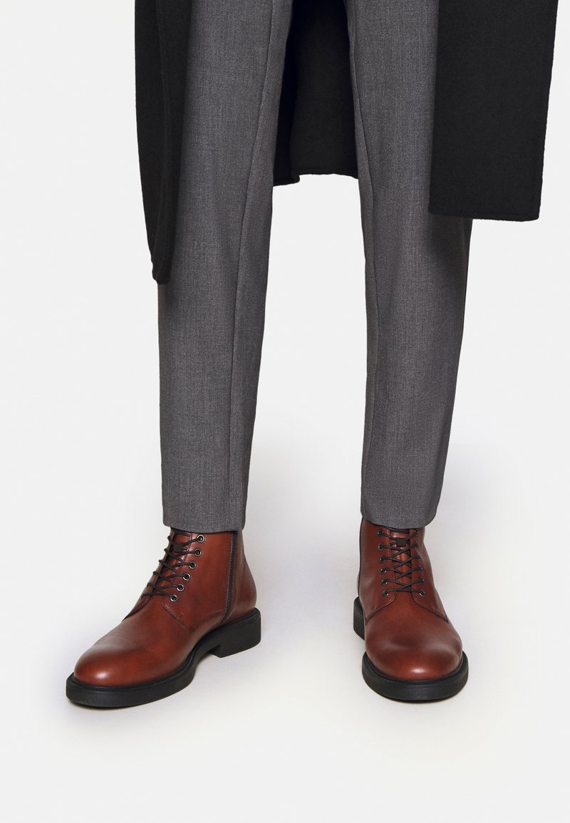 Smeđe kožne gležnjače s okruglim prstima, crnim gumiranim potplatima i vezanjem na rupice. Sive krojene hlače dopunjuju obuću.