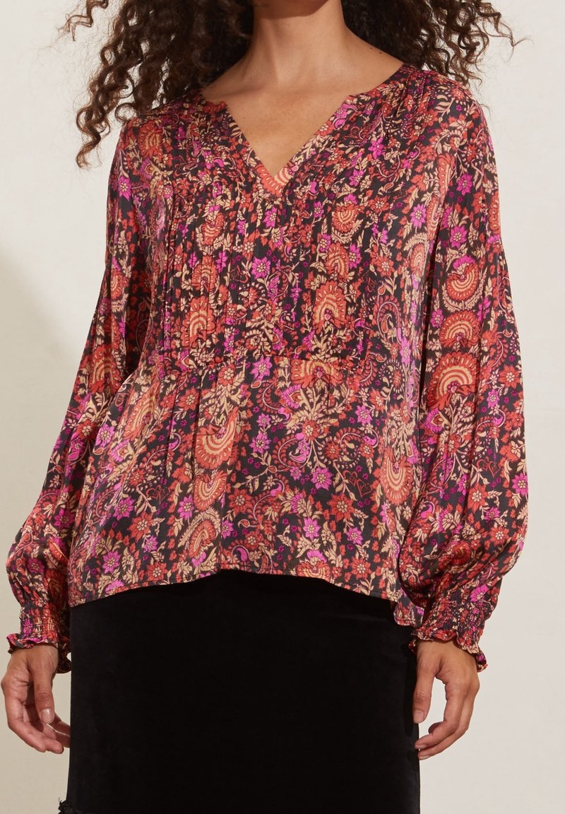 Blouse à motif floral avec manches longues bouffantes et poignets smockés, associée à une jupe noire, portée par une personne aux cheveux bouclés.