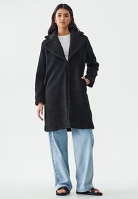 Abrigo de lana negra con cuello muesca y bolsillos frontales, que presenta un tejido texturizado. Combinado con jeans de pierna ancha en un azul claro y sandalias.