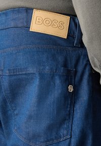 Plave traperice s tamno smešnom kožnom etiketom urezanom s "BOSS", sadrže srebrnu metalnu akcentu i klasični dizajn džepova.