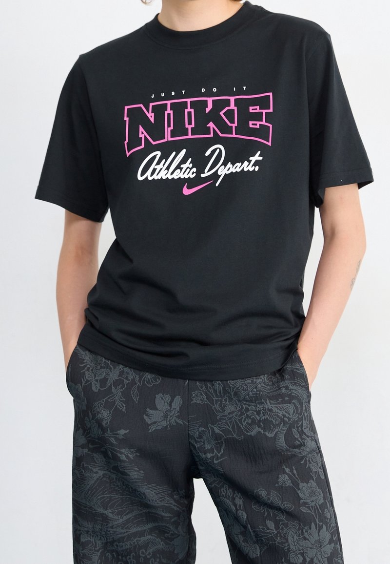 Černé bavlněné tričko s výrazným růžovým a bílým textem „NIKE“ a sloganem „Just Do It“ nahoře. Krátké rukávy a volný střih.