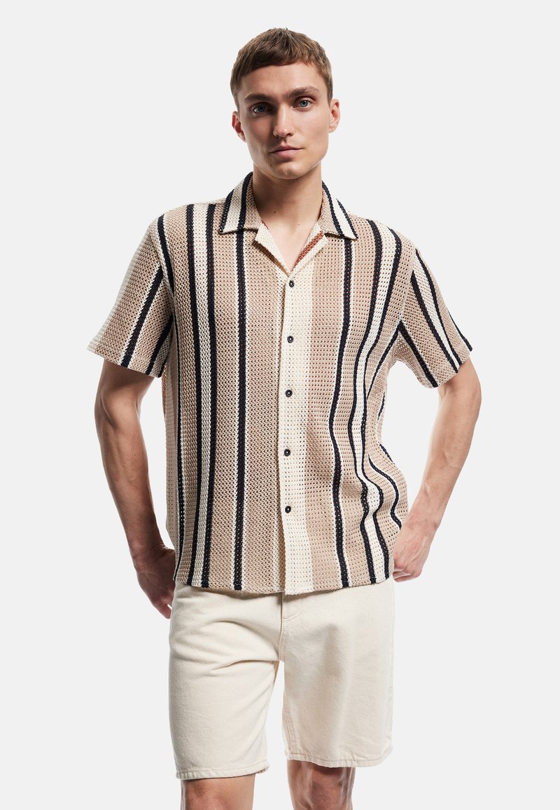 Kortærmet skjorte i en lys beige strik med lodrette marineblå striber, der har en button-down krave og seks sorte knapper. Båret med lyse shorts.