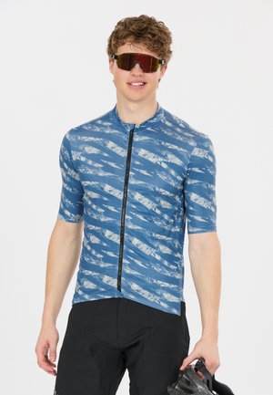 Junger Mann trägt ein blau gemustertes Fahrradtrikot, schwarze Shorts und rot getönte Sportsonnenbrille, hält einen schwarzen Fahrradhelm in der Hand.