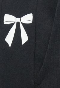 Black fabric hoodie featuring a large white bow graphic on the front. The texture is soft, with ribbed accents near the pockets.