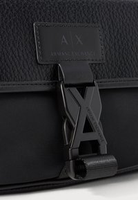 Melna tekstūrēta ādas soma ar matētu melnu aizdari, kurai ir "A|X Armani Exchange" logotips un izšūta zīmola birka virs aizdara.