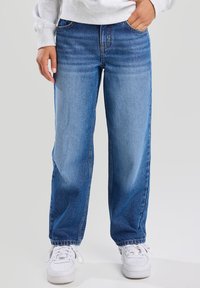 Vaaleansiniset farkut, joissa on rento istuvuus, suora lahjemuoto ja näkyvät saumat. Yhdistetty valkoisiin tennareihin.