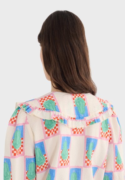 Femme aux cheveux châtain moyen portant un chemisier avec des motifs en forme de poires vertes et de carrés à carreaux rouges, vue de dos sur un fond uni.