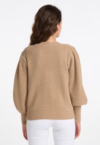 Beige bordás pulóver puffos hosszú ujjakkal, textúrált anyaggal és kerek nyakkivágással; szűk fehér nadrággal párosítva.