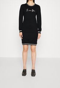 Zwarte jurk met lange mouwen en witte logotekst, met een contrasterende gestreepte kraag en manchetten, en een rechte silhouet. Draagt zwarte mules met blokhak.