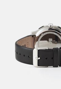 Bracciale in pelle nera con fibbia in metallo argentato, dotato di fori punzonati per la regolazione. Include un logo del marchio sulla fibbia.