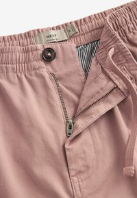 Pantalon rose coupe droite avec taille élastique, fermeture éclair partiellement ouverte, boutonnière et doublure intérieure rayée visible à la taille.