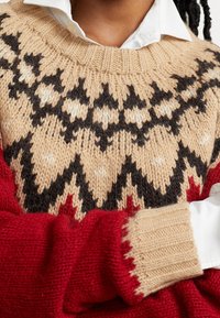 Pull en tricot présentant un corps beige avec un motif géométrique noir et crème, des manches rouges et des poignets côtelés. Fabriqué en fil texturé.