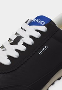 Baskets noires à lacets blancs, marque "HUGO" sur le côté et languette bleue, gros plan montrant le tissu texturé et les détails de la couture.
